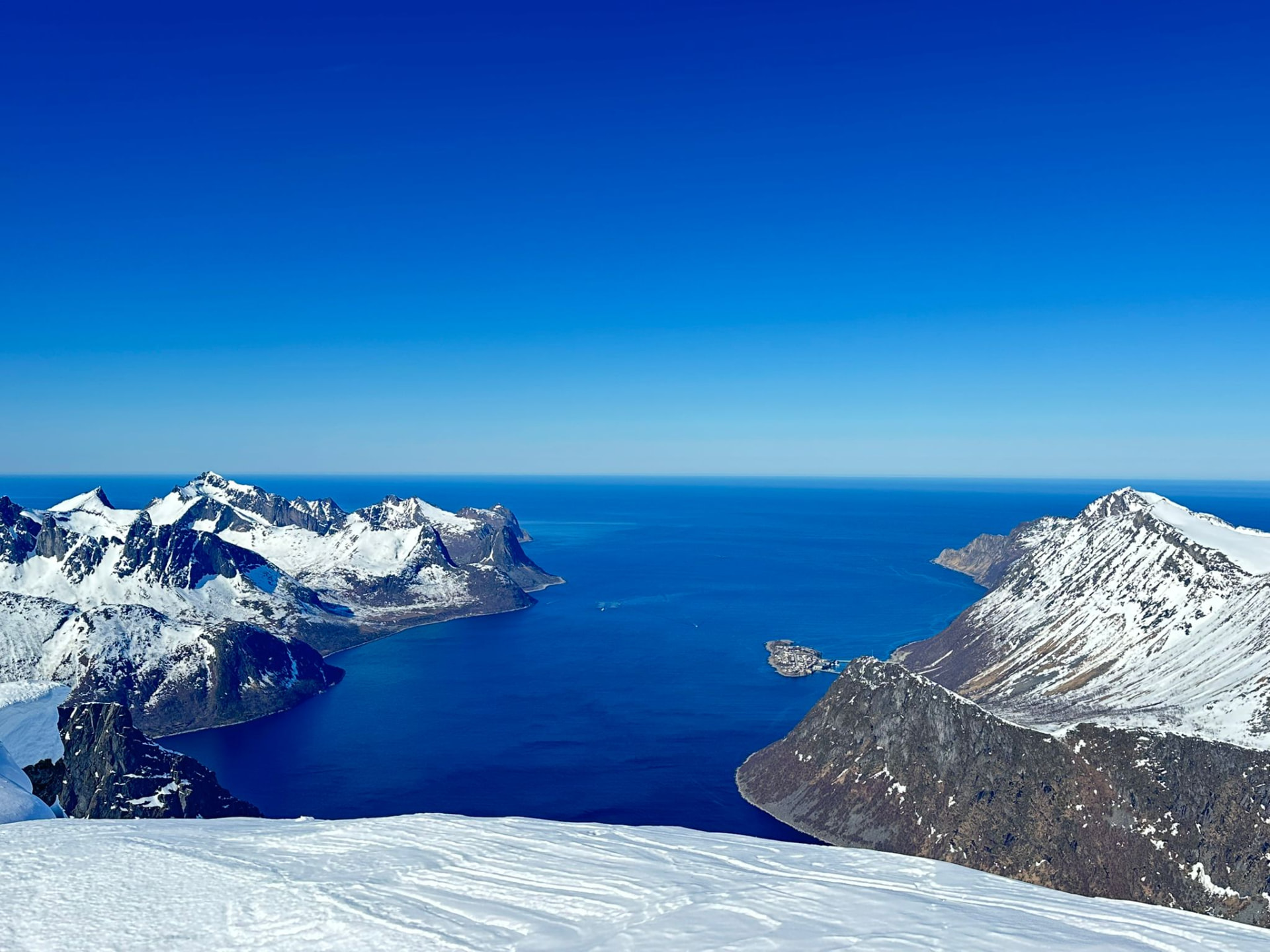 Utsikt från en snötäckt topp över djupblå fjord och snöklädda toppar